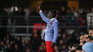 Un supporter enthousiaste, vêtu d'un blazer bleu et d'un pantalon rouge, gesticule avec passion lors d'un match de football marocain, entouré de spectateurs surexcités.