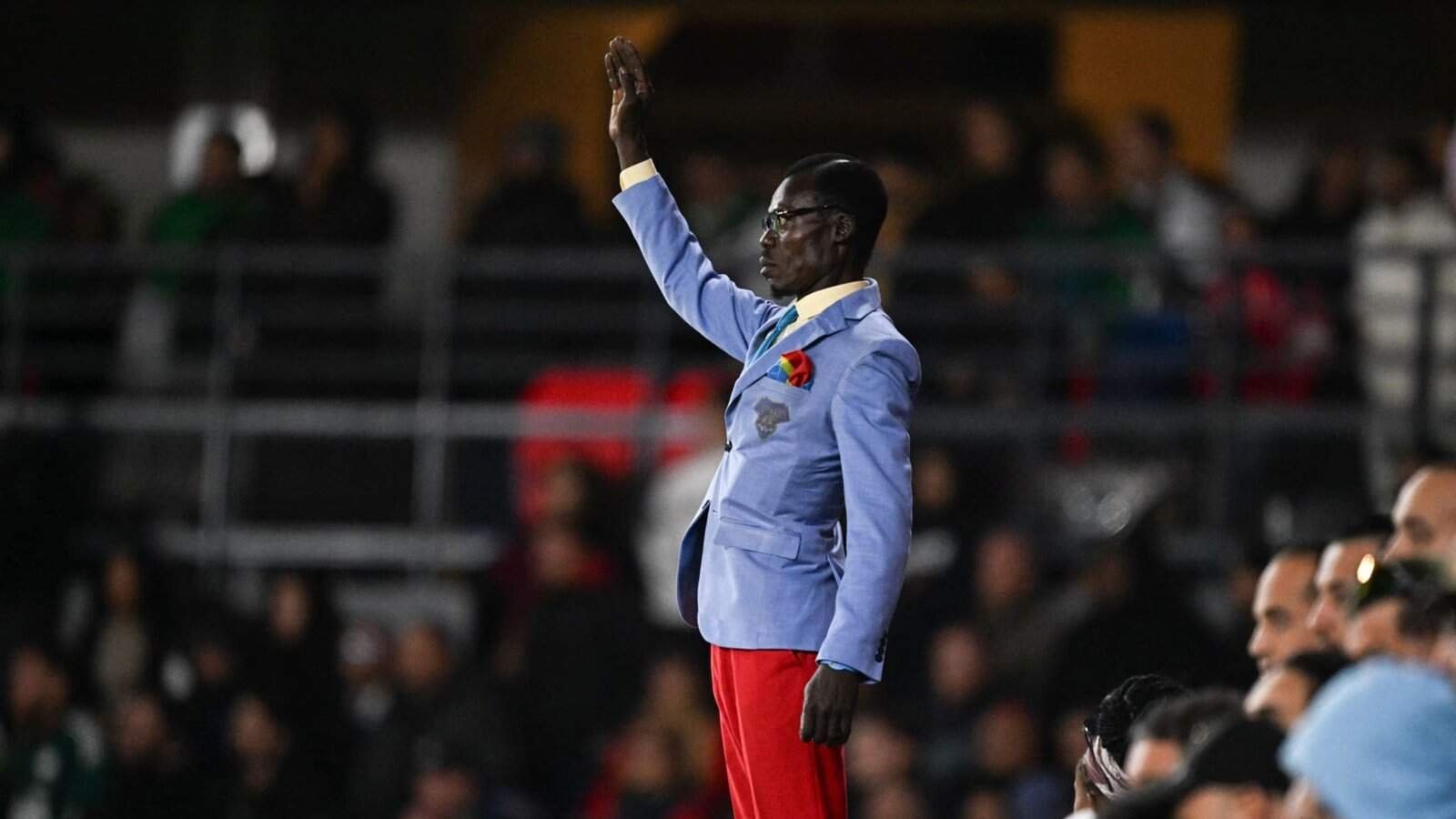 Un supporter enthousiaste, vêtu d'un blazer bleu et d'un pantalon rouge, gesticule avec passion lors d'un match de football marocain, entouré de spectateurs surexcités.