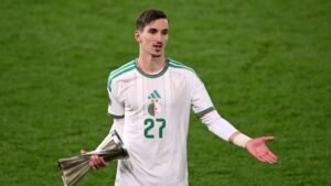 Joueur algérien portant le maillot numéro 27, en tenue blanche avec des accents verts, tenant un trophée et gesticulant sur le terrain.