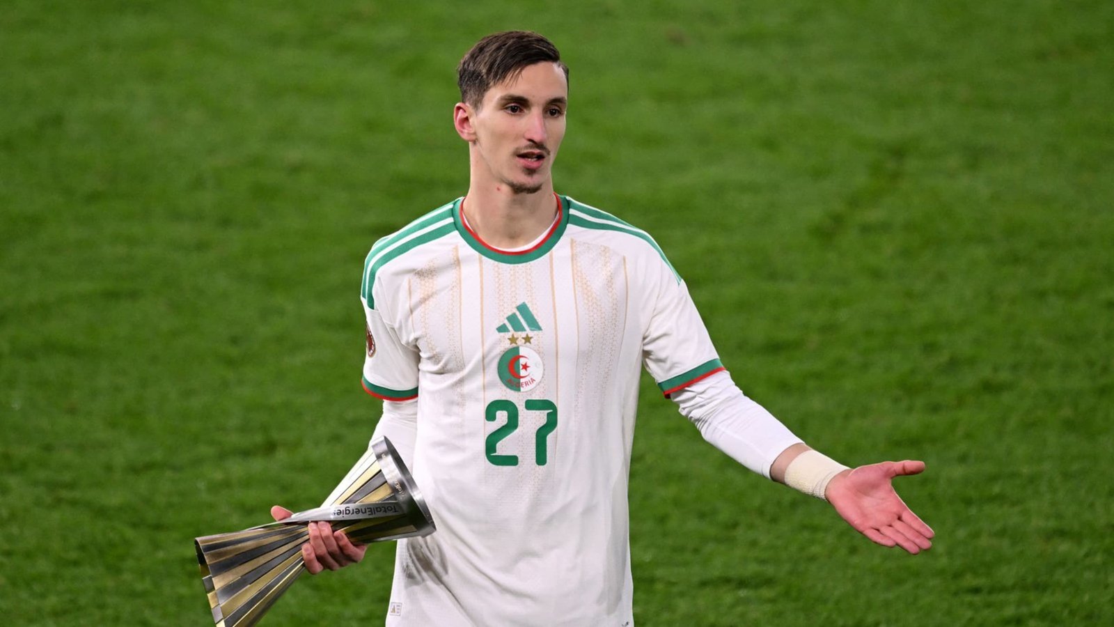 Joueur algérien portant le maillot numéro 27, en tenue blanche avec des accents verts, tenant un trophée et gesticulant sur le terrain.