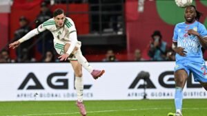 A Moroccan player in a white and green kit attempts a shot on goal, while a defender in a blue kit prepares to intercept.