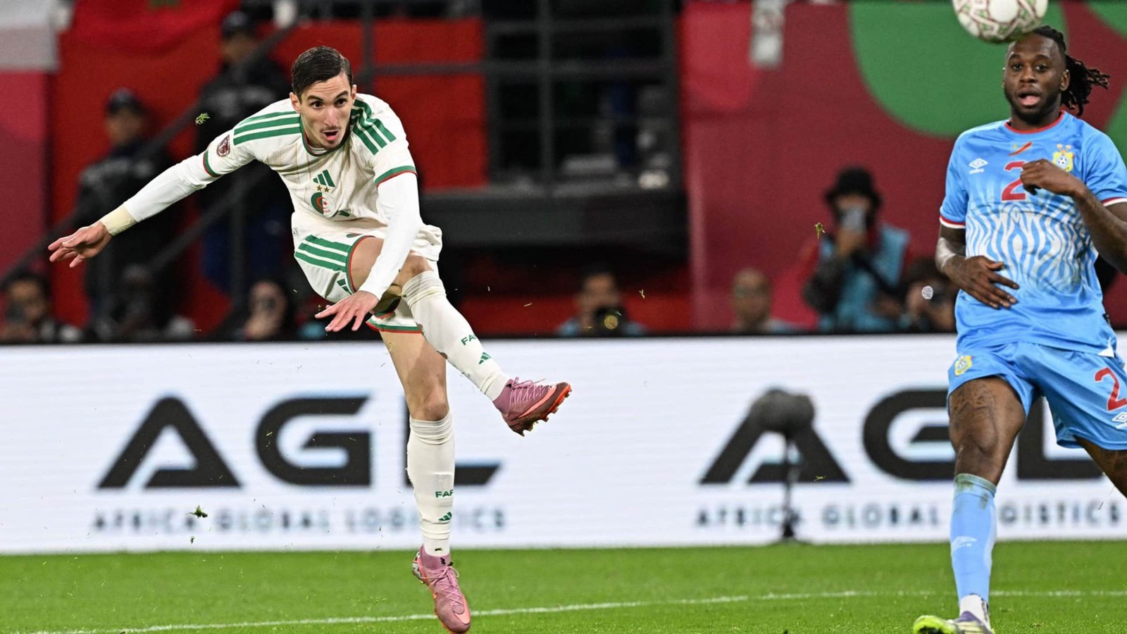 A Moroccan player in a white and green kit attempts a shot on goal, while a defender in a blue kit prepares to intercept.