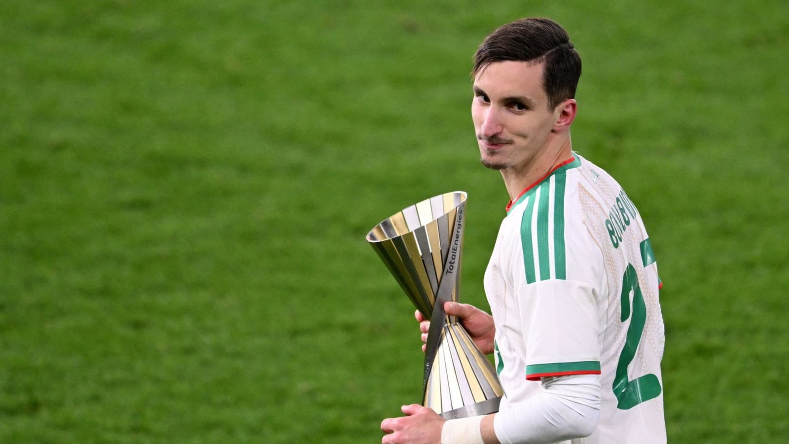 Boufal sourit en tenant le trophée du Championnat d'Afrique des Nations TotalEnergies, vêtu de l'uniforme blanc et vert du Maroc sur le terrain.