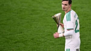 Joueur algérien tenant un trophée en argent sur le terrain, portant un maillot blanc avec des accents verts, célébrant la victoire après un match.