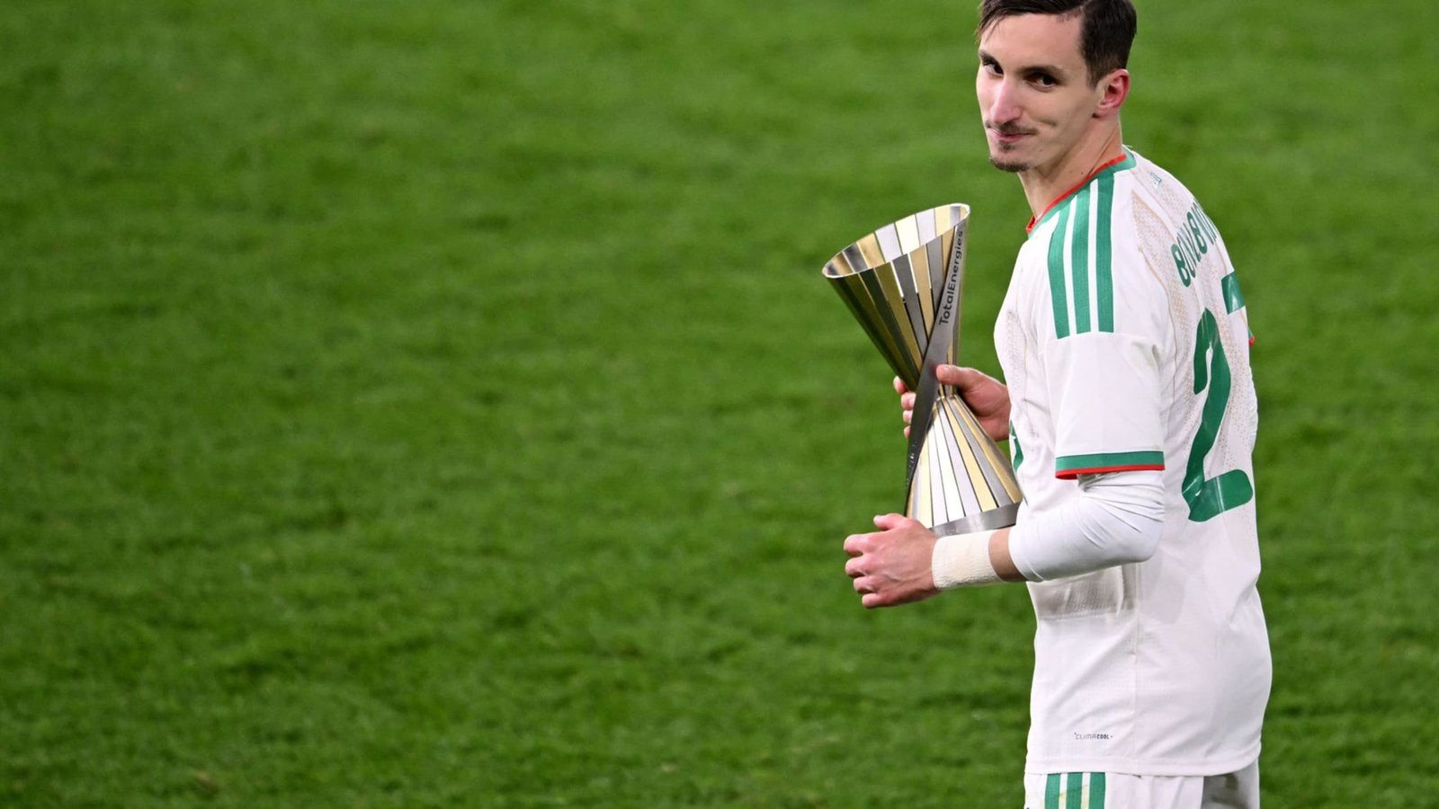 Joueur algérien tenant un trophée en argent sur le terrain, portant un maillot blanc avec des accents verts, célébrant la victoire après un match.