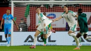 Les joueurs algériens célèbrent un but, vêtus de leurs tenues vert et blanc, tandis qu'un joueur adverse observe à l'arrière-plan.