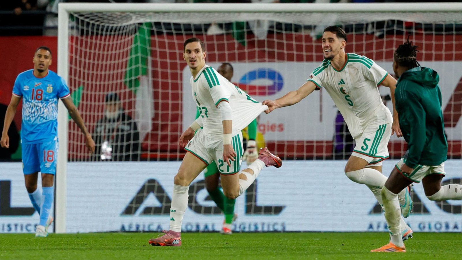 Des joueurs algériens célèbrent un but pendant un match, l'un d'eux sprintant joyeusement tandis qu'un autre observe. Le but est visible en arrière-plan.