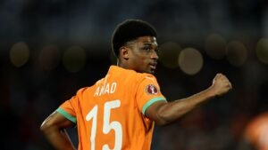 Amad Diallo, wearing an orange national team jersey with the number 15, celebrates, with a blurred crowd in the background.