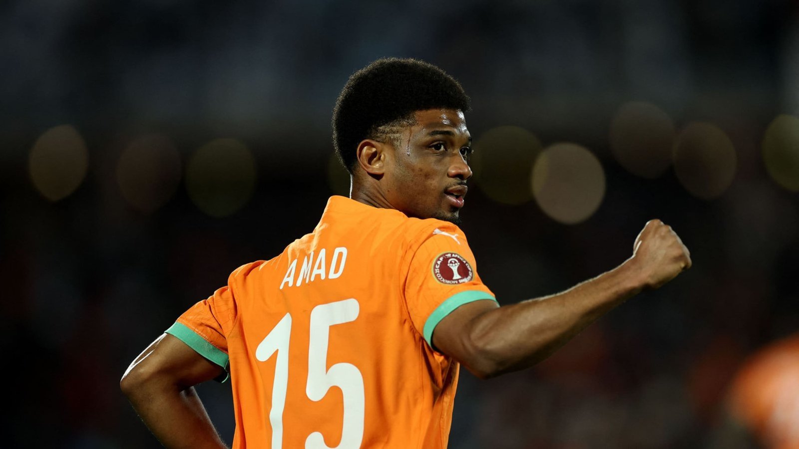 Amad Diallo, wearing an orange national team jersey with the number 15, celebrates, with a blurred crowd in the background.