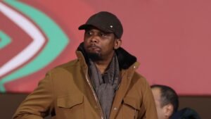 Samuel Eto'o regarde le match avec attention, portant un manteau marron et une casquette noire sur un fond rouge et vert vif.