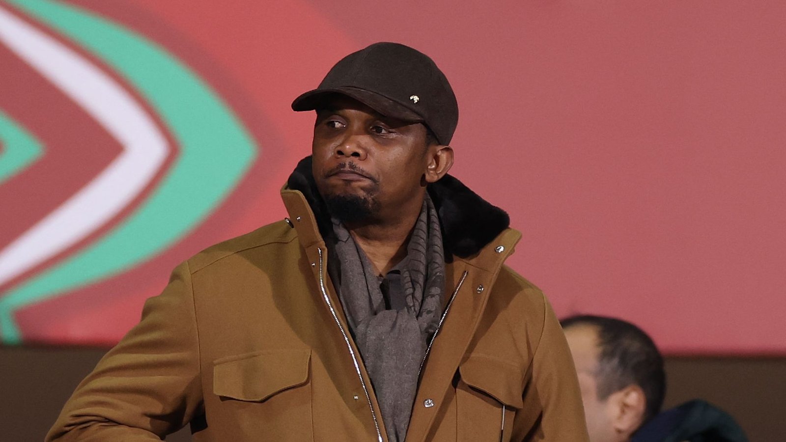 Samuel Eto'o regarde le match avec attention, portant un manteau marron et une casquette noire sur un fond rouge et vert vif.