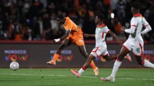Ivorian player in an orange kit attempts a shot while Moroccan defenders in white and red close in during a competitive match.