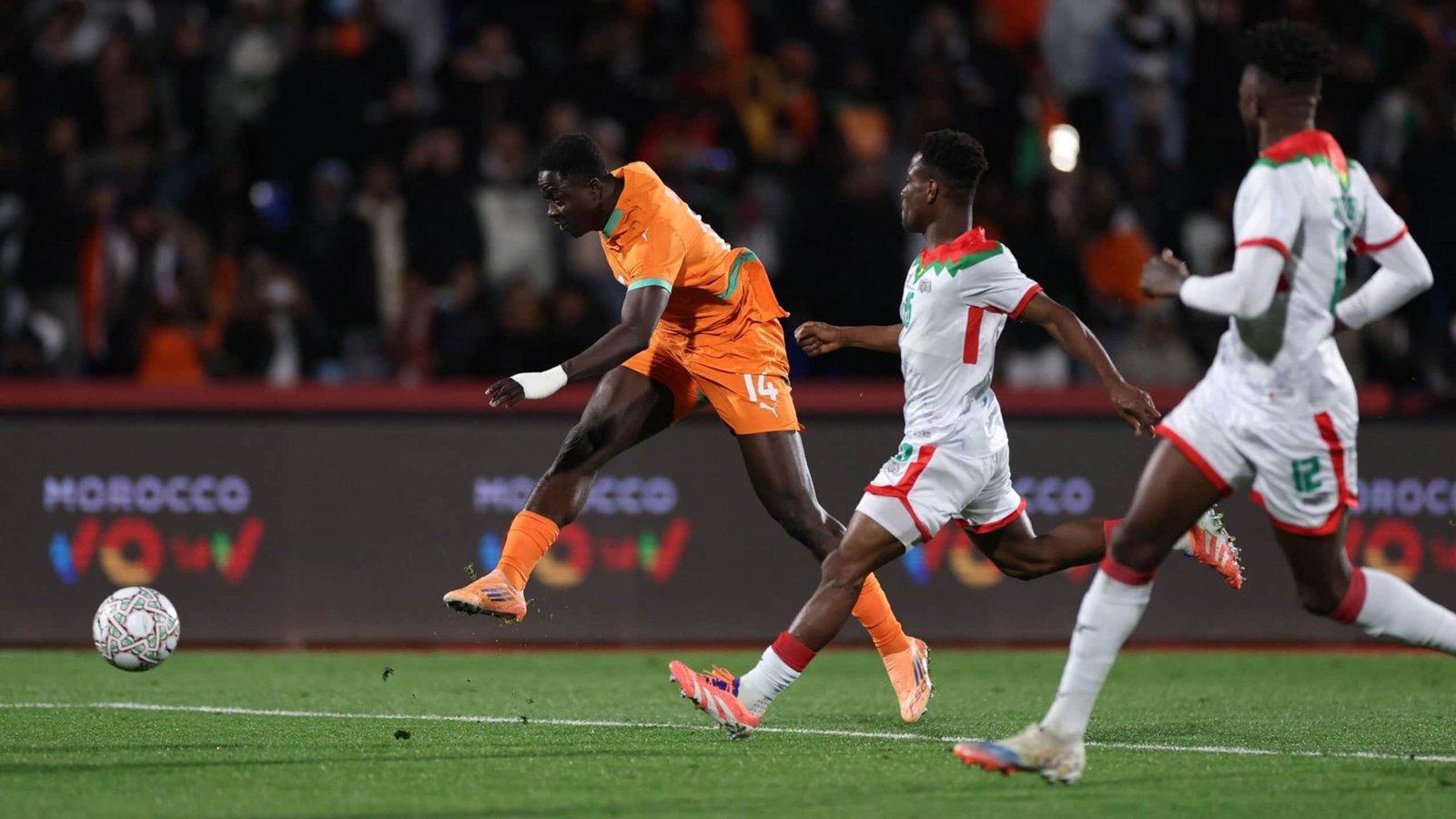 Ivorian player in an orange kit attempts a shot while Moroccan defenders in white and red close in during a competitive match.