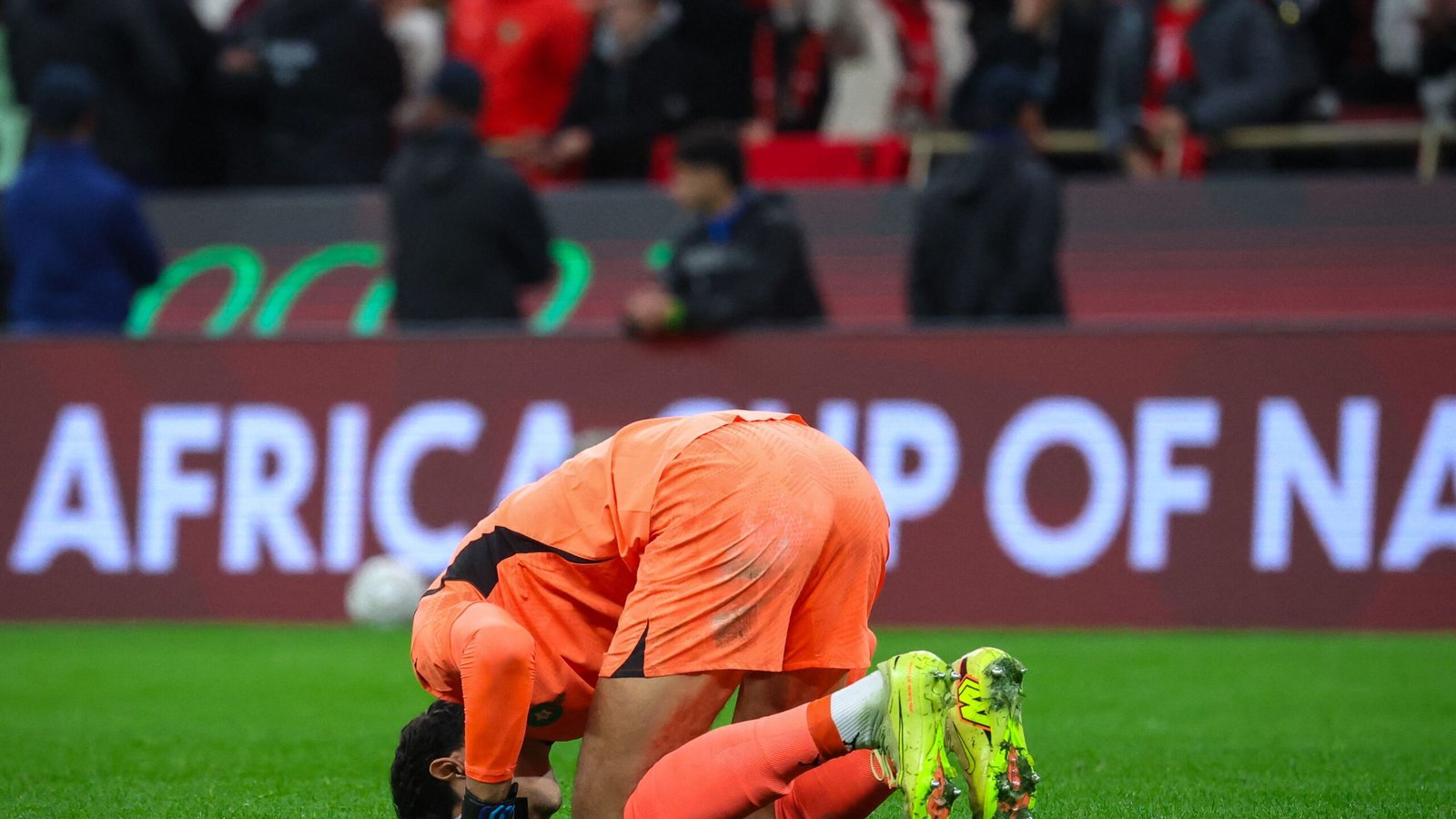 Moroccan goalkeeper kneeling on the pitch, visibly emotional, with "AFRICA CUP OF NATIONS" displayed prominently in the background.