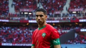Achraf Hakimi, portant le maillot rouge du Maroc avec un col vert, semble concentré sur le terrain, avec une foule vibrante en rouge derrière lui.