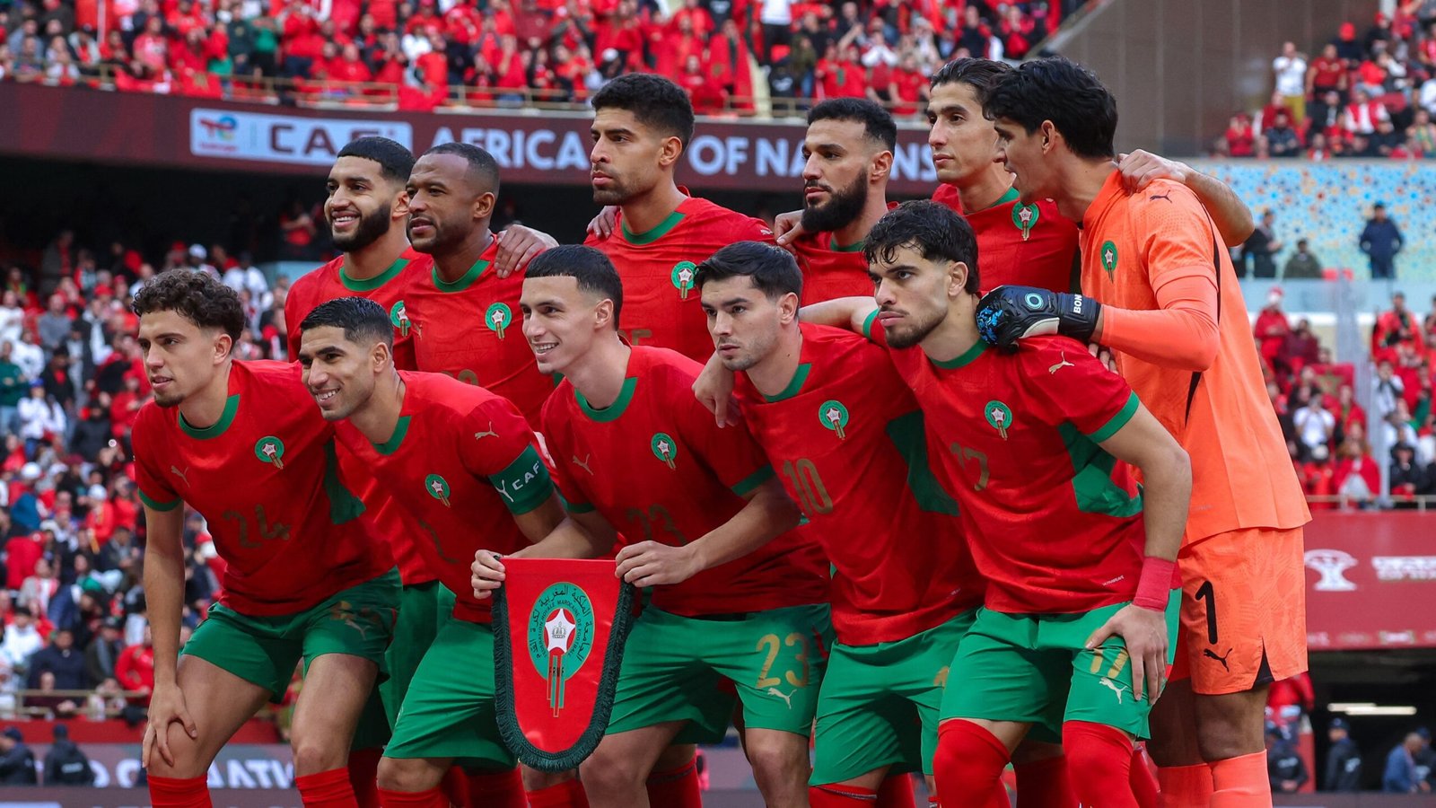 L'équipe nationale du Maroc pose pour une photo, portant des kits rouges avec des garnitures vertes, tenant fièrement un bouclier avant un match de l'AFCON.