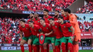 L'équipe nationale de football du Maroc pose avec assurance dans ses tenues rouges éclatantes, avec des supporters en arrière-plan, avant un match.