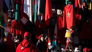 Moroccan fans in red attire showcase national pride amidst a vibrant display of flags and merchandise in a bustling market setting.
