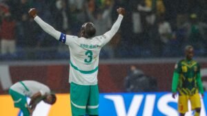 Koulibaly célèbre sous la pluie, portant le maillot vert et blanc du Sénégal, avec un joueur dégonflé en arrière-plan lors d'un match tendu.
