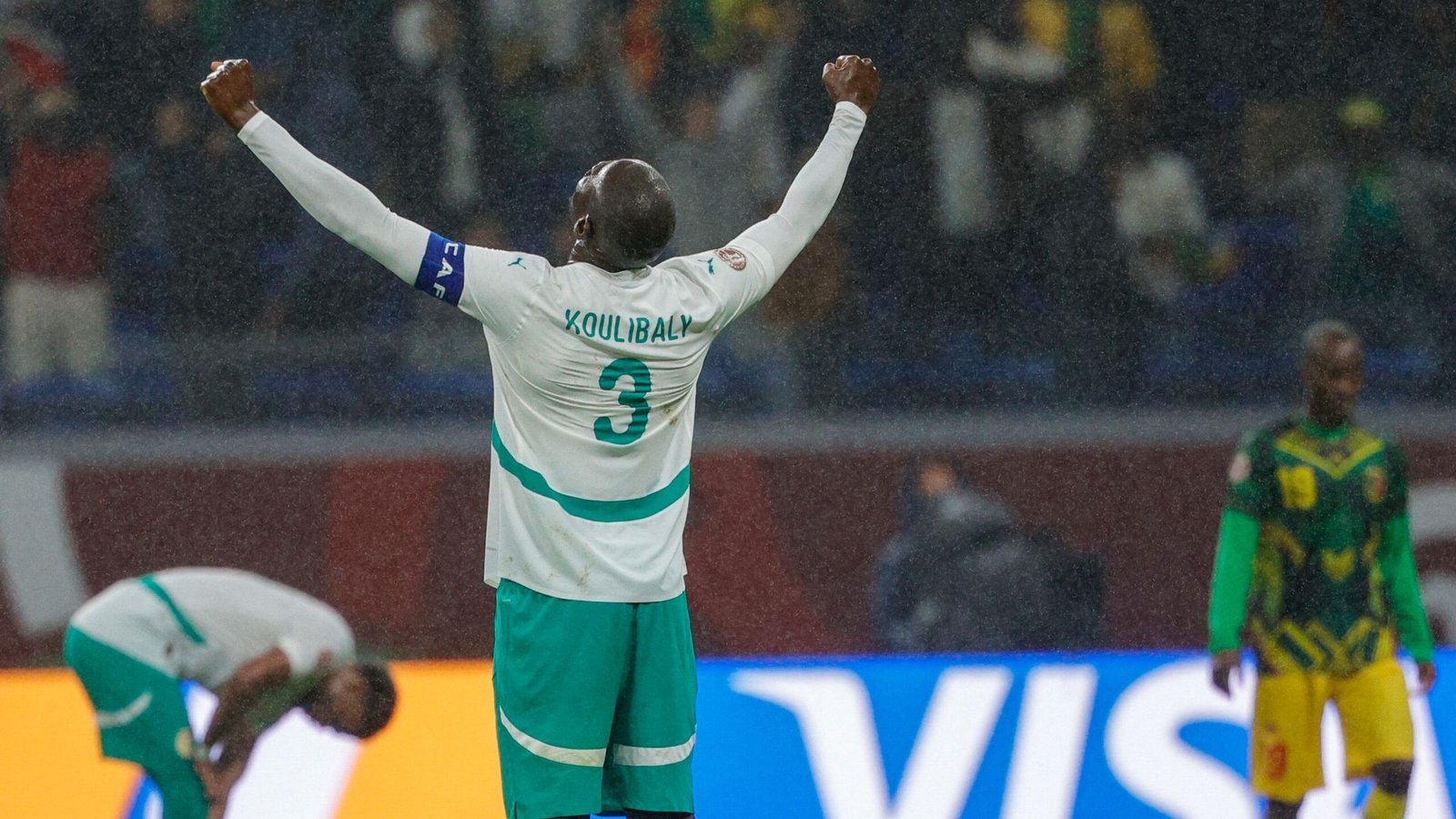Koulibaly célèbre sous la pluie, portant le maillot vert et blanc du Sénégal, avec un joueur dégonflé en arrière-plan lors d'un match tendu.