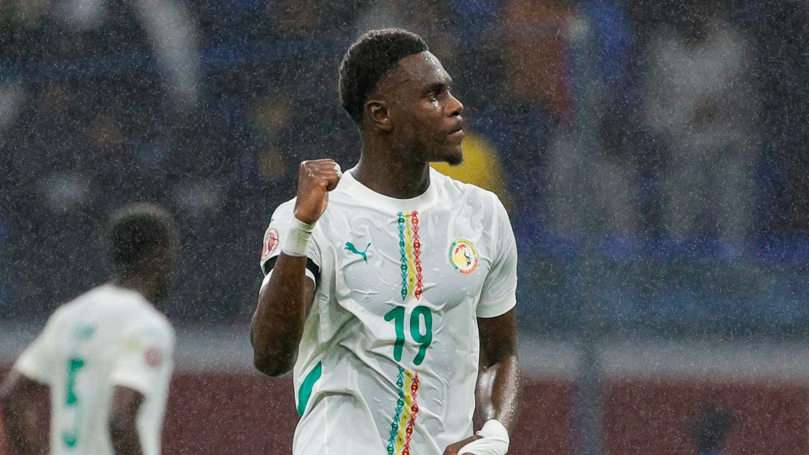 Le joueur sénégalais célèbre avec un poing serré, portant un maillot blanc avec des détails verts et rouges, lors d'une scène de match pluvieuse.