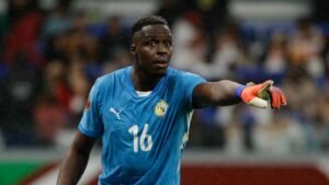 Le gardien de but sénégalais portant un maillot bleu avec le numéro 16, gesticulant pendant un match, avec des supporters flous en arrière-plan.