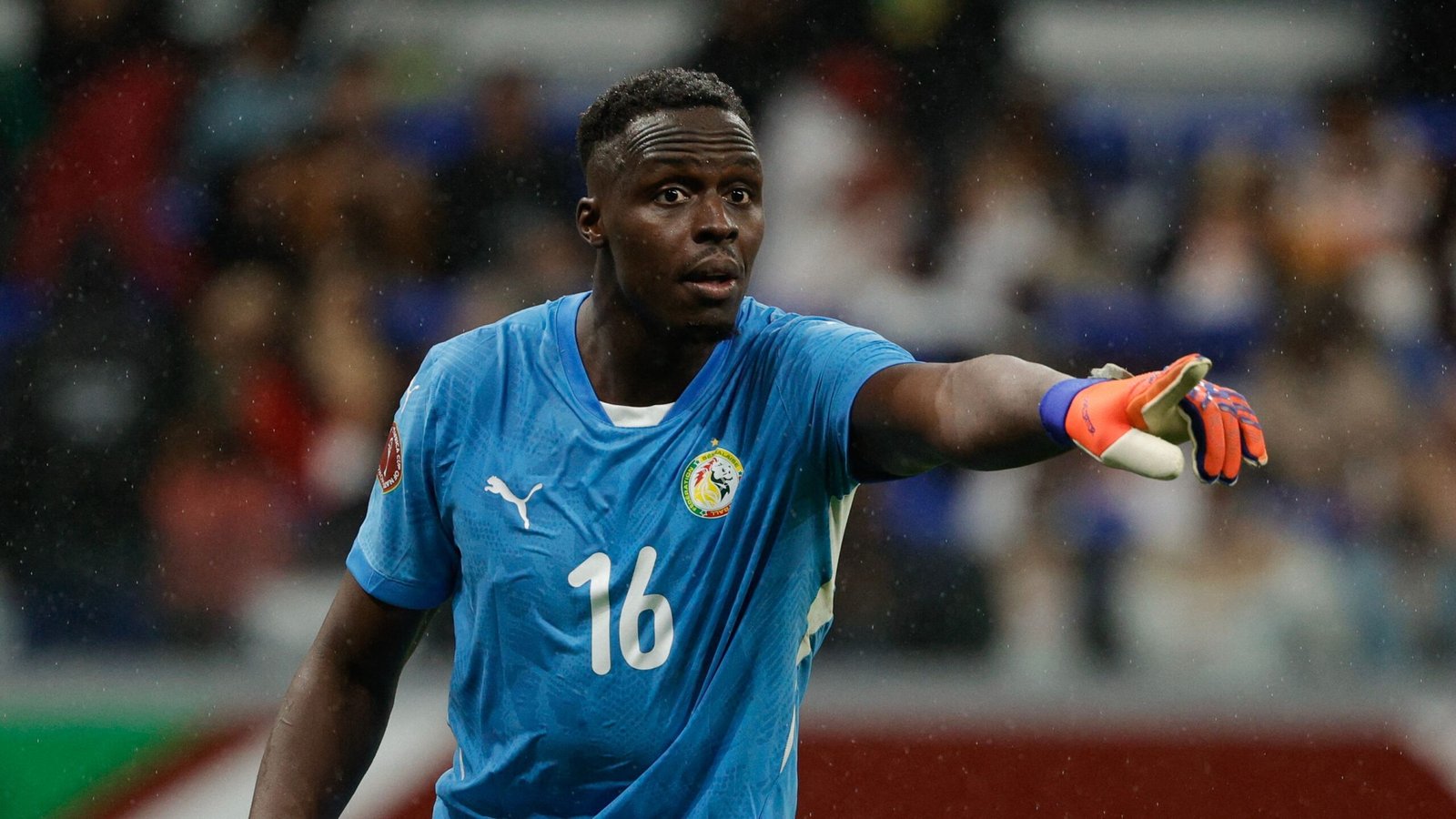 Le gardien de but sénégalais portant un maillot bleu avec le numéro 16, gesticulant pendant un match, avec des supporters flous en arrière-plan.