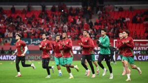 Les joueurs de l'équipe nationale du Maroc s'échauffent en maillots rouges et verts avant un match, avec une foule vibrante visible en arrière-plan.