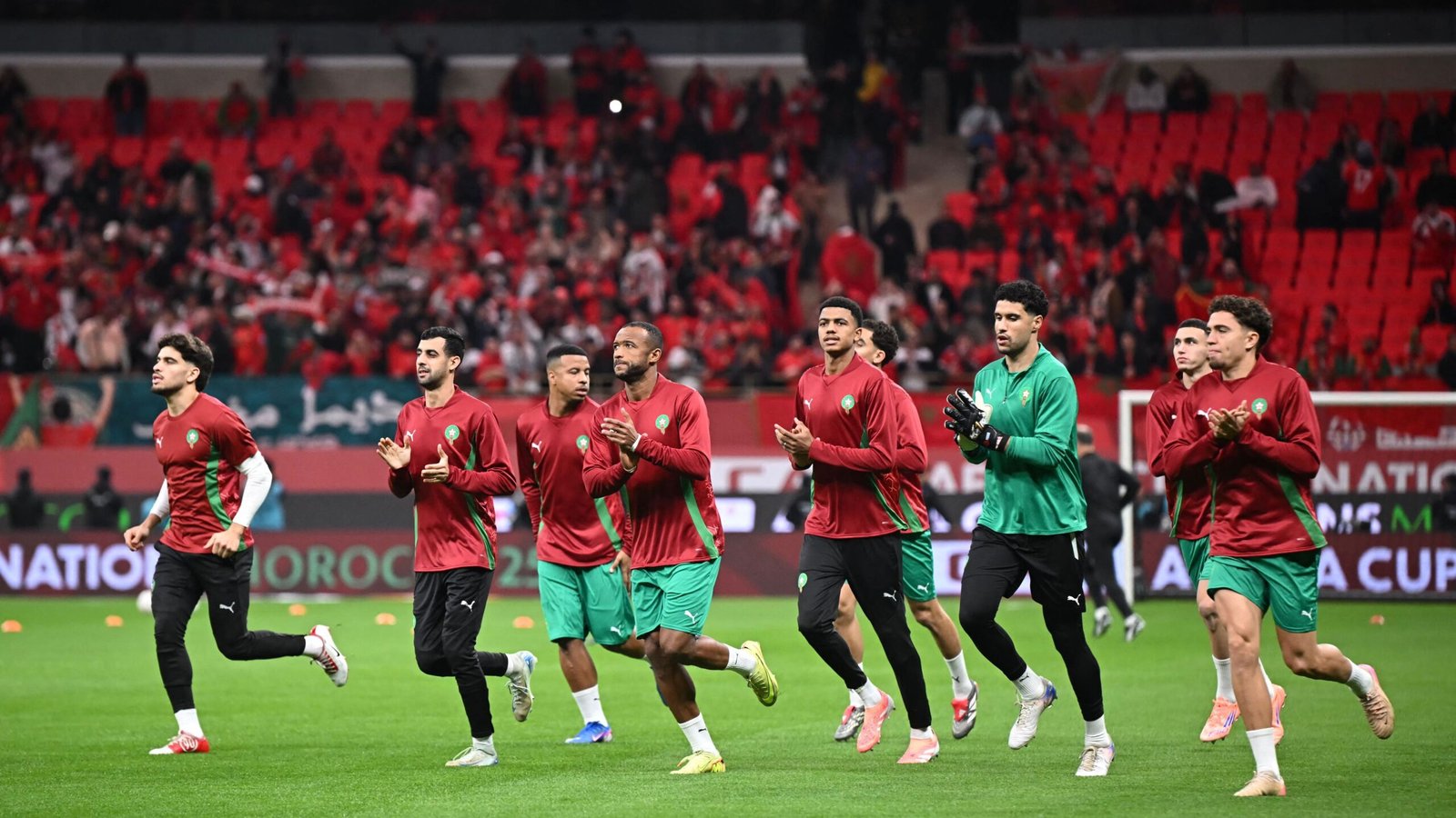 Les joueurs de l'équipe nationale du Maroc s'échauffent en maillots rouges et verts avant un match, avec une foule vibrante visible en arrière-plan.