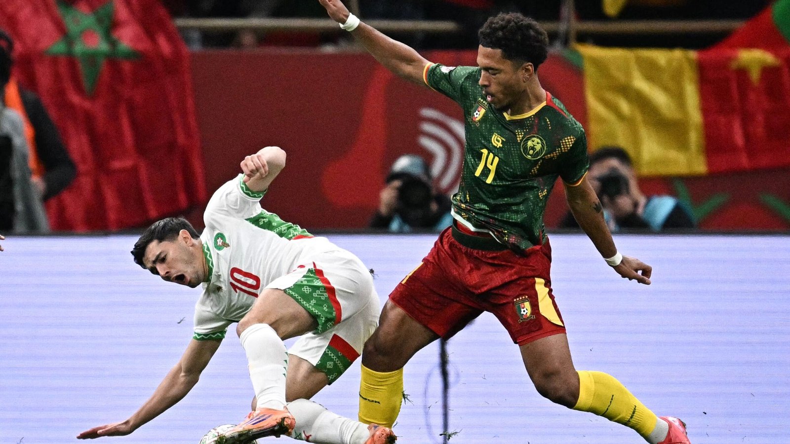 Le joueur marocain #10 tombe alors qu'il est défié par le camerounais #14 lors d'un match tendu, avec des drapeaux visibles en arrière-plan.