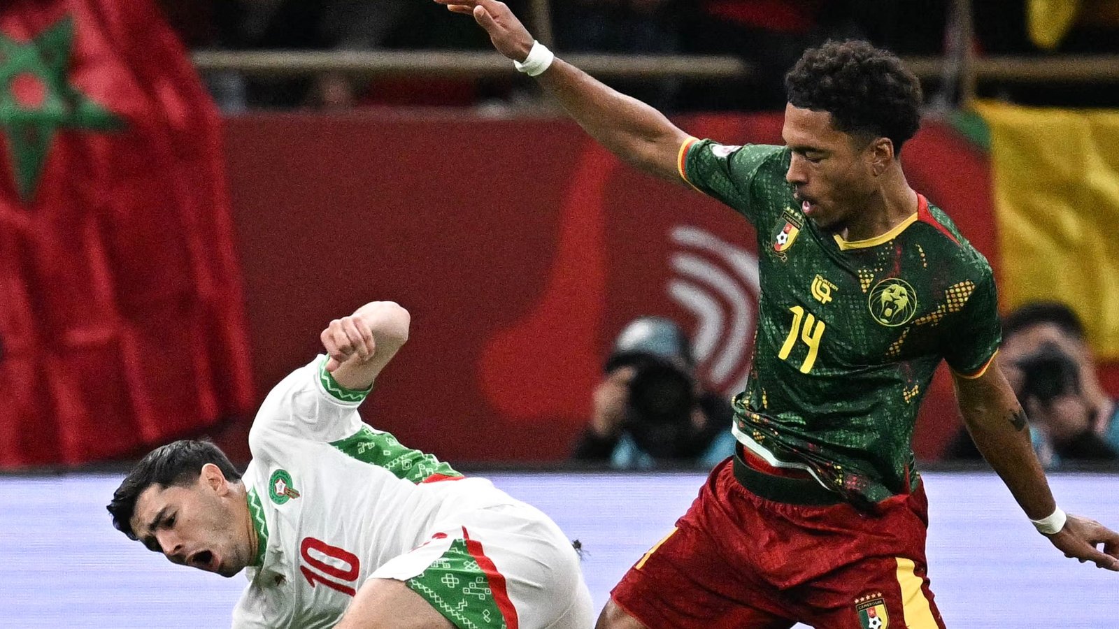 Le joueur marocain #10 tombe au sol alors que le #14 du Cameroun proteste lors d'un match international tendu, avec des drapeaux vibrants en arrière-plan.
