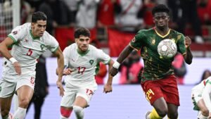 Les défenseurs marocains poursuivent un joueur camerounais (n° 24) contrôlant le ballon lors d'un match intense, mettant en valeur des maillots d'équipe vibrants.