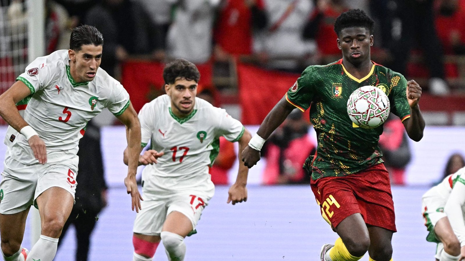 Les défenseurs marocains poursuivent un joueur camerounais (n° 24) contrôlant le ballon lors d'un match intense, mettant en valeur des maillots d'équipe vibrants.