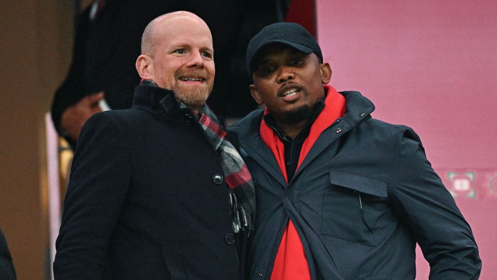 Two men smile and pose together, one in a black coat with a patterned scarf, the other in a navy jacket with a red collar, against a pink backdrop.