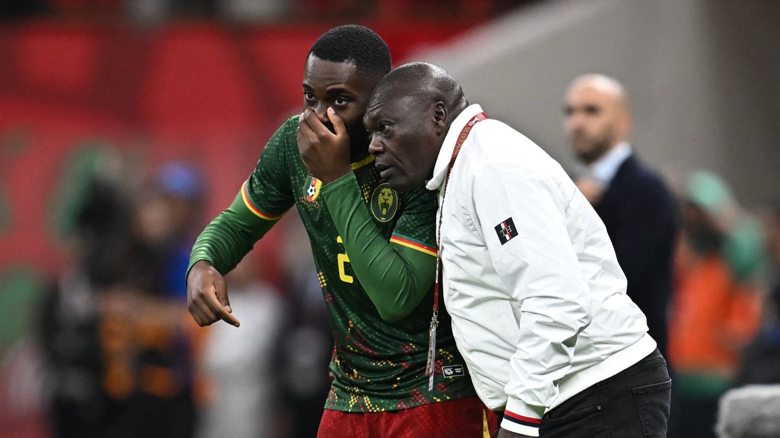 Cameroonian players discussing strategy; one in a green kit with red and yellow accents, the other in a white jacket, late-match tension.