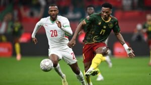 Un joueur marocain portant le numéro 20 poursuit un joueur camerounais dans leur tenue verte et jaune vibrante lors d'une action de match intense.