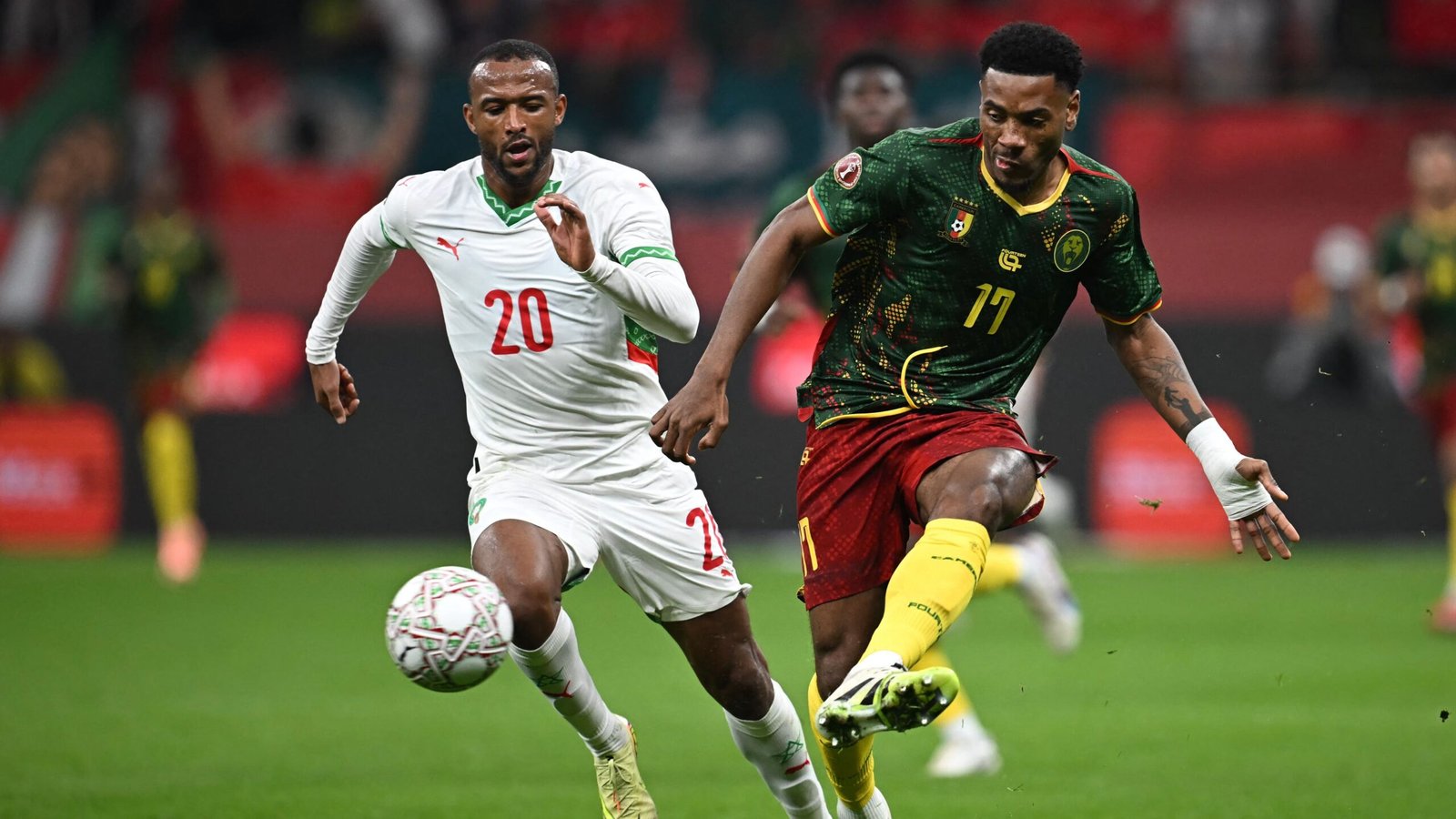 Un joueur marocain portant le numéro 20 poursuit un joueur camerounais dans leur tenue verte et jaune vibrante lors d'une action de match intense.