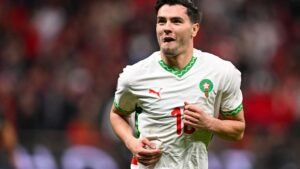 Le joueur marocain, portant un maillot blanc rehaussé de vert et de rouge, célèbre avec passion après avoir marqué un but lors d'un match.