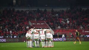 L'équipe de football du Maroc se regroupe sur le terrain, vêtue de blanc et de vert, au milieu de fans en rouge acclamant lors d'un match.
