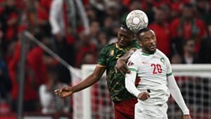 A Moroccan player, wearing a white kit with green accents, grimaces as he clashes with a Cameroonian opponent in a dramatic match moment.