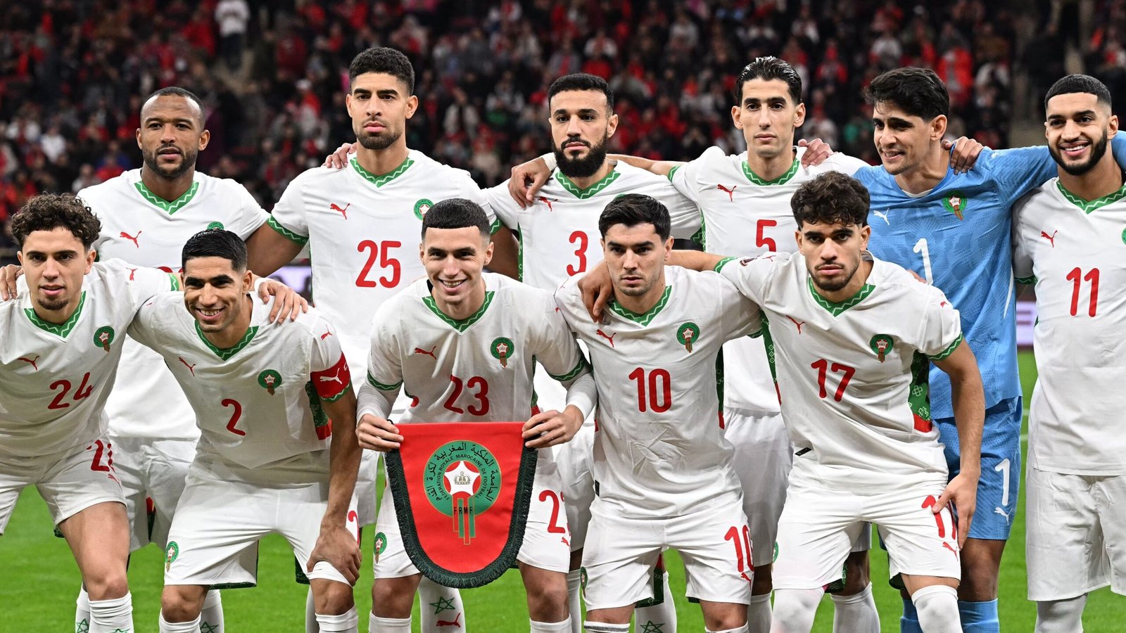 L'équipe nationale de football du Maroc pose pour une photo de groupe, portant des maillots blancs avec des accents verts, tenant un emblème de bouclier avant un match.