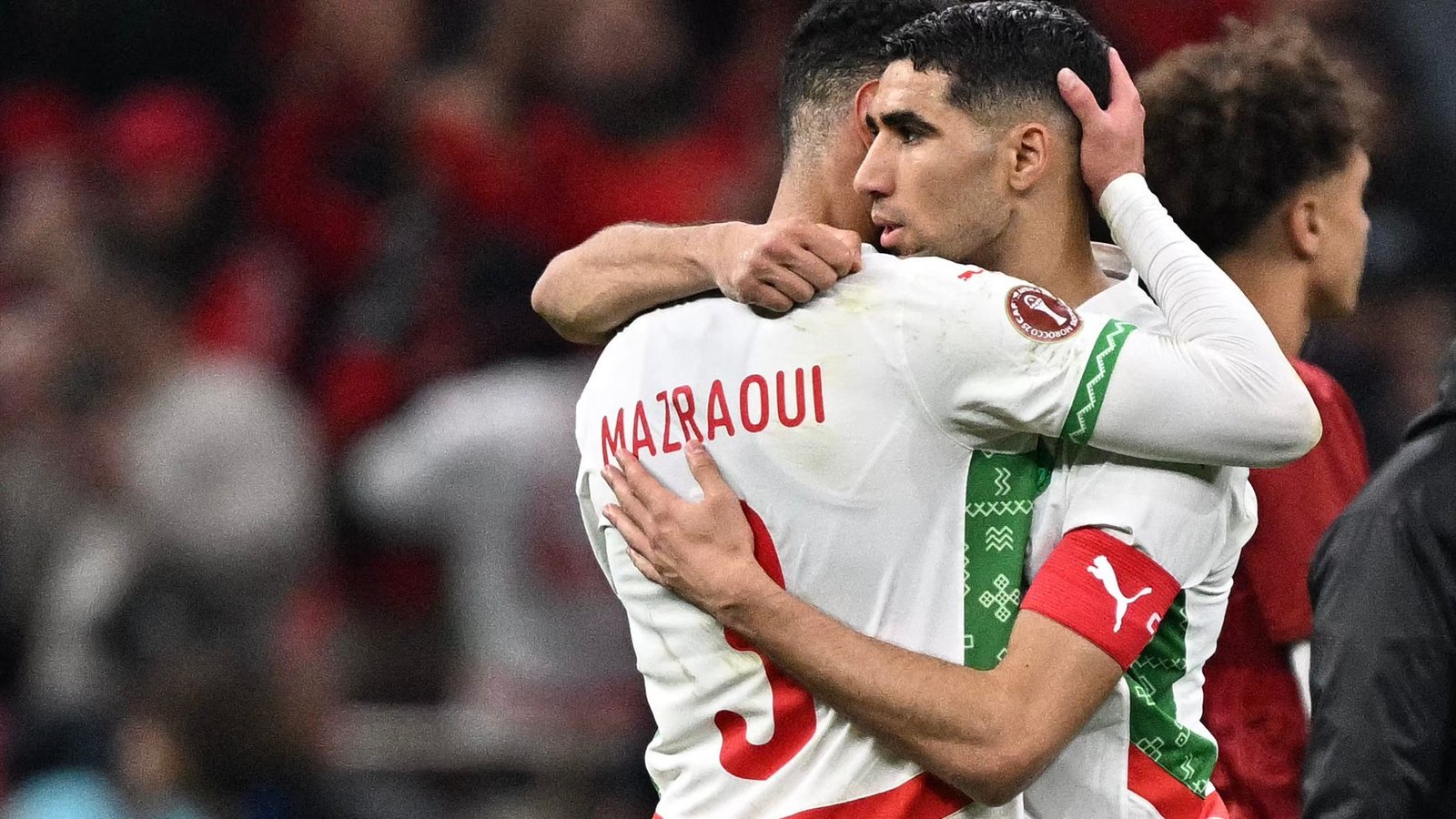 Achraf Hakimi embrasse Noussair Mazraoui après un match, tous deux portant les maillots de l'équipe nationale marocaine avec des accents rouges et verts, montrant de la camaraderie.