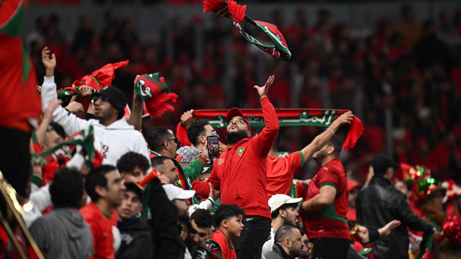Moroccan fans celebrate passionately in vibrant red and green attire, waving scarves and cheering in a packed stadium atmosphere.