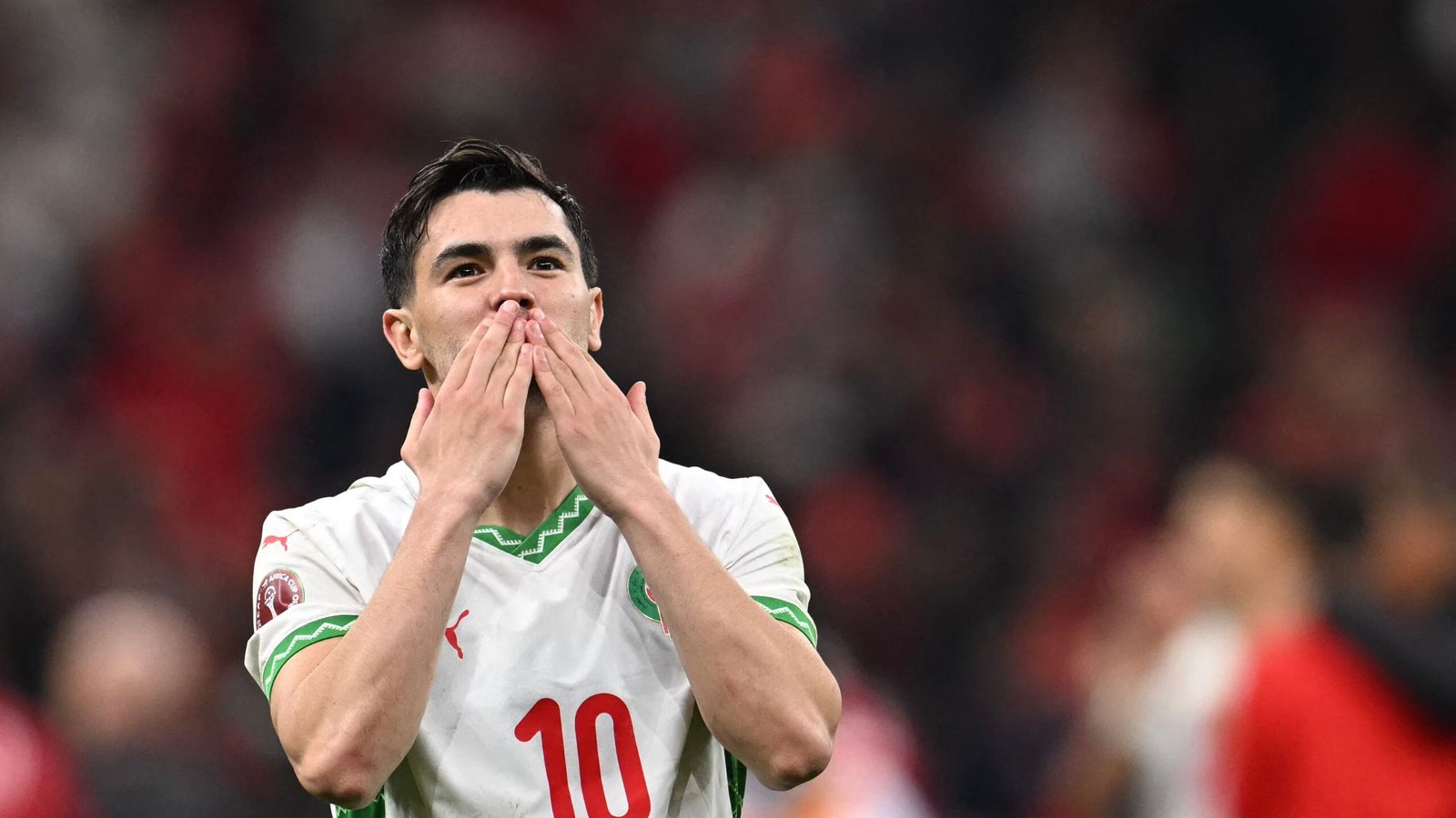 Moroccan player celebrating with a kiss gesture, wearing the national team's white kit with green accents, amidst a cheering crowd.