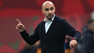 Walid Regragui gesticule avec passion en bord de touche, portant une veste noire sur une chemise claire, se détachant sur un fond rouge vif.