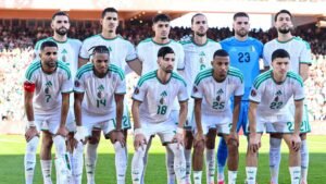L'équipe nationale de football algérienne pose pour une photo d'équipe, portant des maillots blancs avec des détails verts, avant un match dans un stade vibrant.