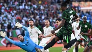 Le joueur nigérian, portant un maillot vert, saute pour tirer pendant que le gardien algérien plonge, avec des coéquipiers en action et des fans en arrière-plan.