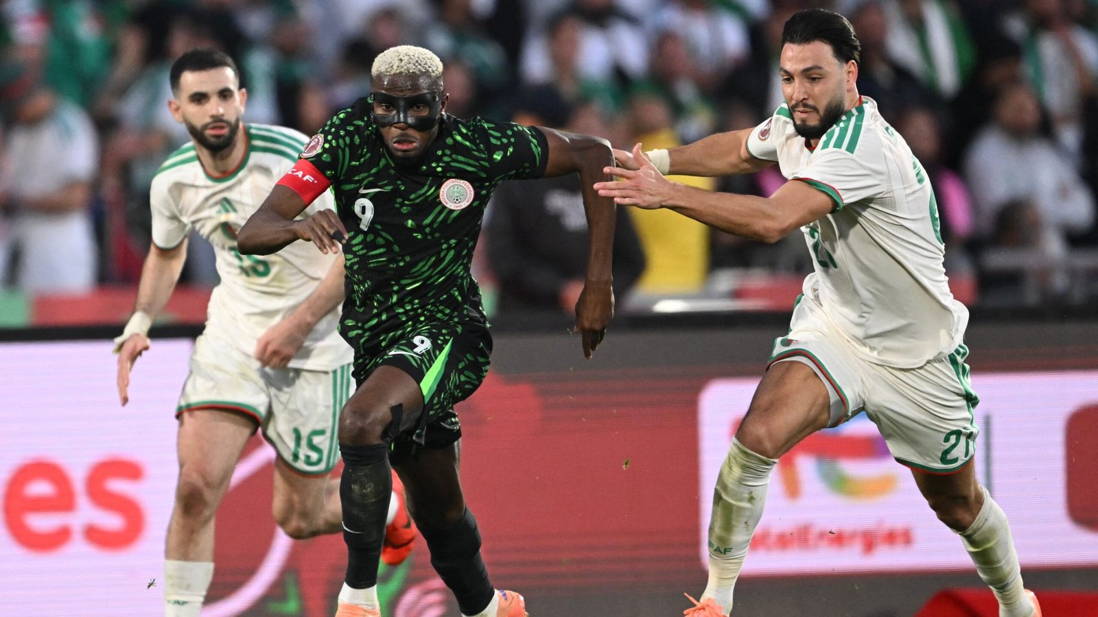 Victor Osimhen, dans un maillot vert du Nigeria, dépasse deux défenseurs algériens lors d'un match intense, montrant une action de football dynamique.
