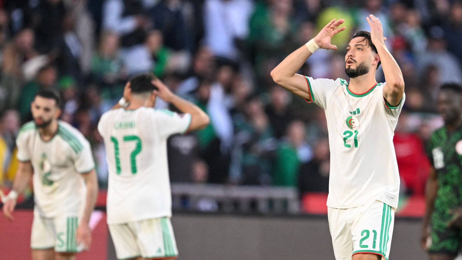Algerian players look dejected during a match, with one player (number 21) raising his arms in frustration, wearing a white kit with green accents.