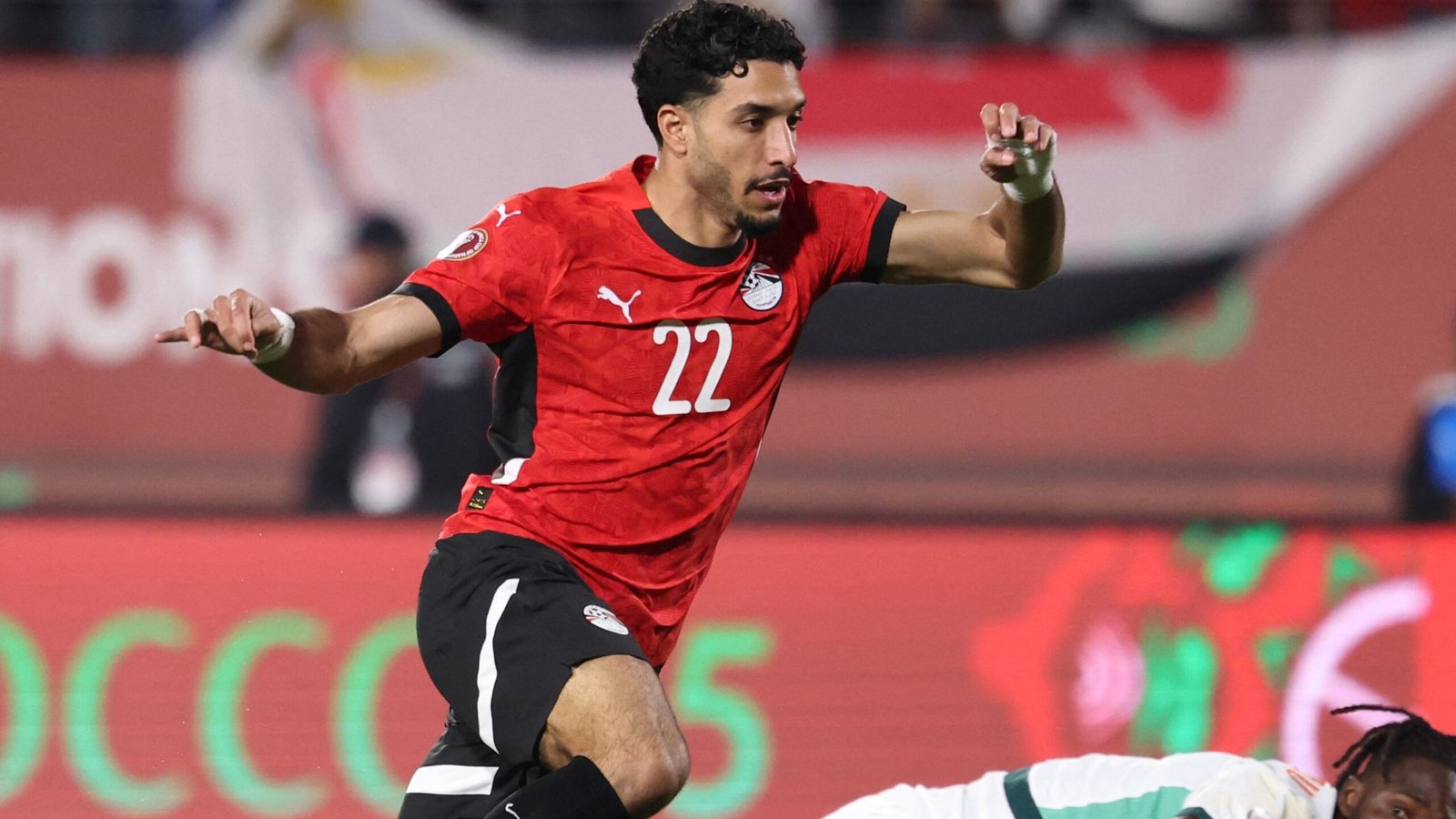 Joueur égyptien en maillot rouge, numéro 22, dribblant lors d'un match contre l'équipe marocaine, montrant une compétition intense et un mouvement dynamique.